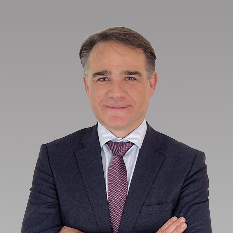 Juan Luis Merino is well-dressed in a black suit, standing with crossed arms.